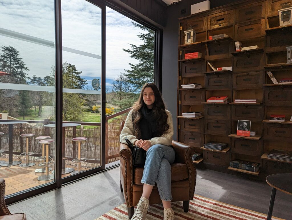 Photo de moi assise pendant ma pratique sur un fauteuil devant une bibliothèque de livres et un paysage calme