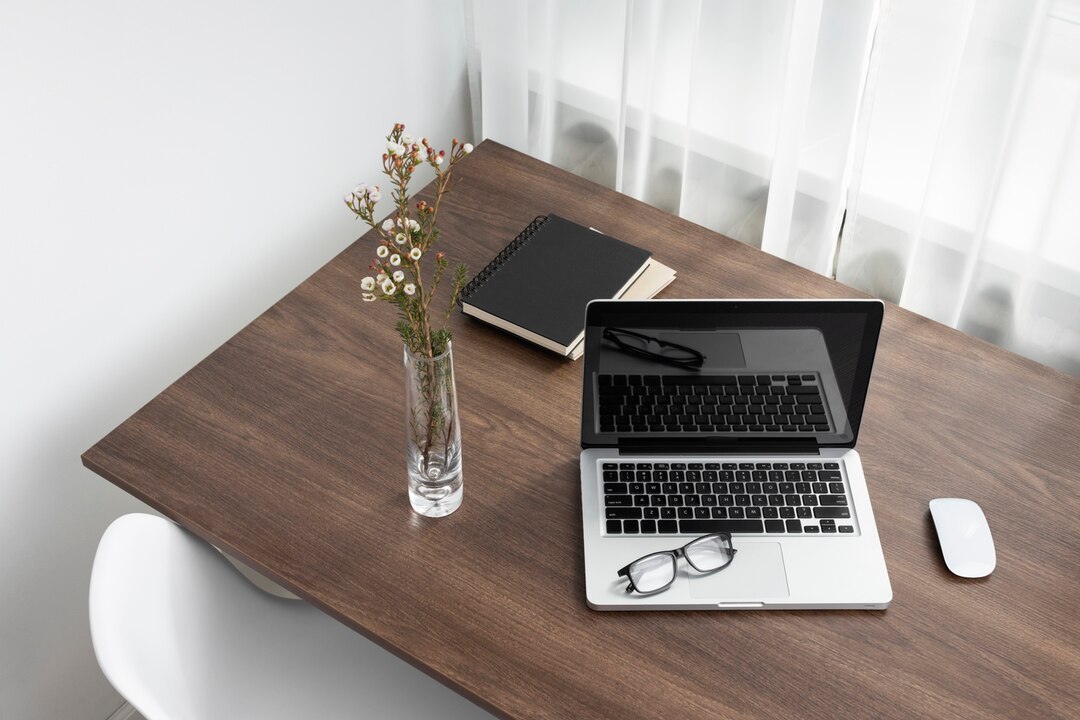 Bureau en bois avec sur le dessus un ordinateur portable, une souris sans fil, des lunettes, un carnet de notes et un bouquet de felurs